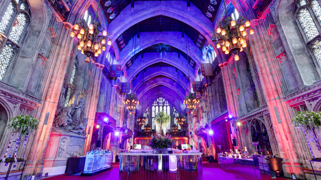 An interior of an elegant, gothic style room with an arched roof, set up for an event with tables and purple lights.